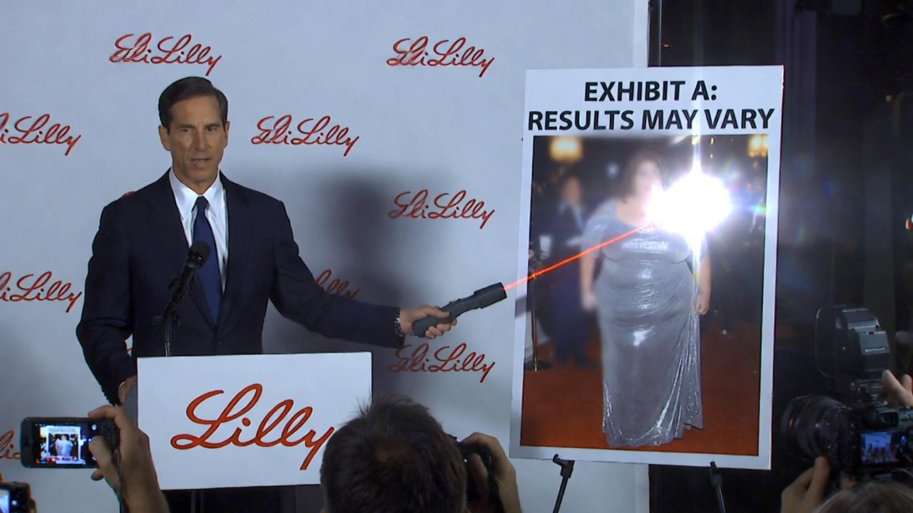 An Eli Lilly spokesperson aims a laser pointer at a laminated red carpet photo labeled Exhibit A.